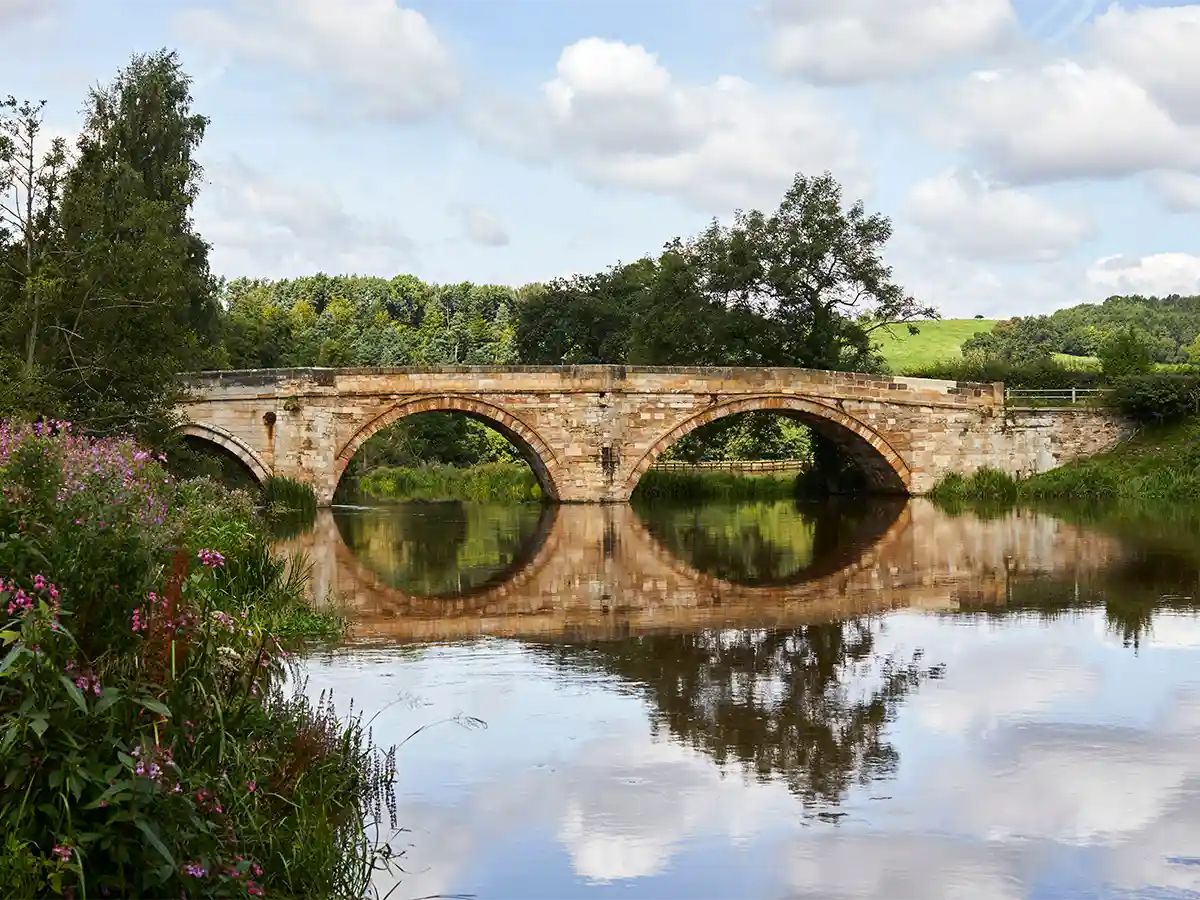 Historic stone bridge over a calm river in East Yorkshire, surrounded by lush greenery and blooming wildflowers on a partly cloudy day.