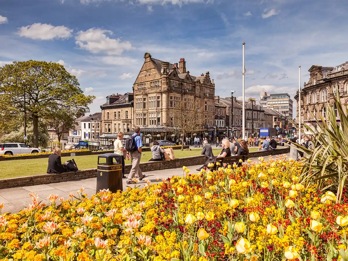 Sunny day in Harrogate, North Yorkshire with people enjoying the park, vibrant flower beds in the foreground, and historic buildings in the background.